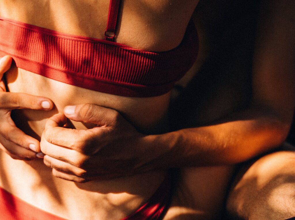 The image shows a close-up of a woman at a swinger party in a red bralette, with hands clasped around the midsection, highlighting their abdominal muscles under warm lighting.