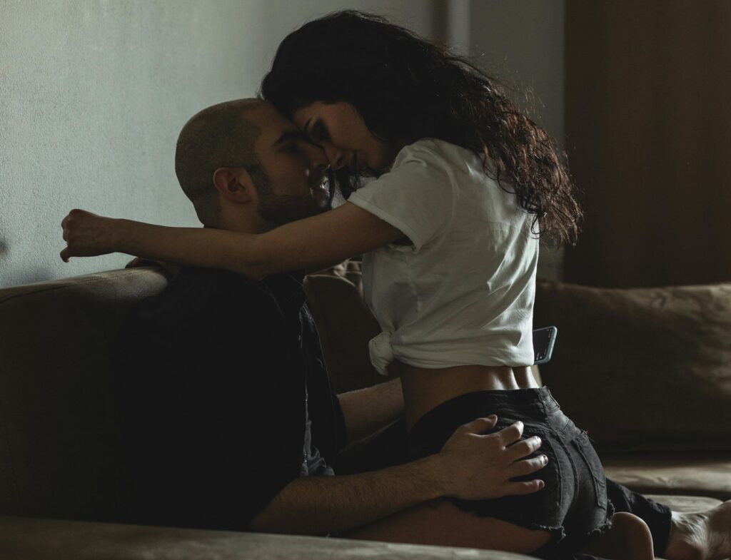 The image depicts an affectionate moment between two swingers embracing each other on a couch in a dimly lit room.