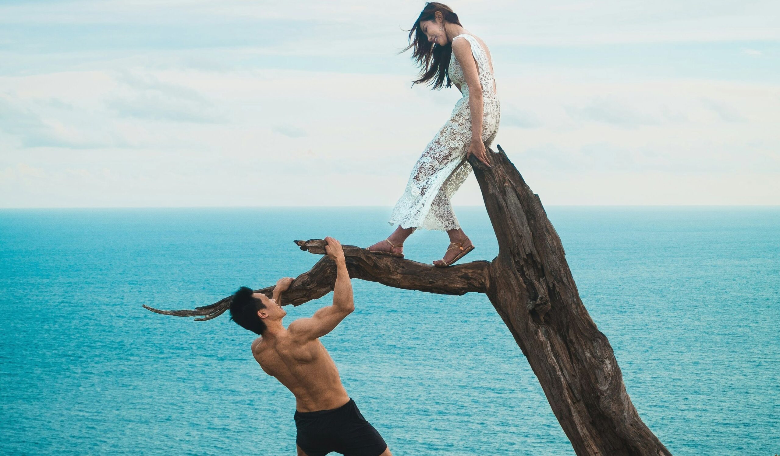 woman sitting on tree trunk with man holding on branch near sea under white clouds during daytime
