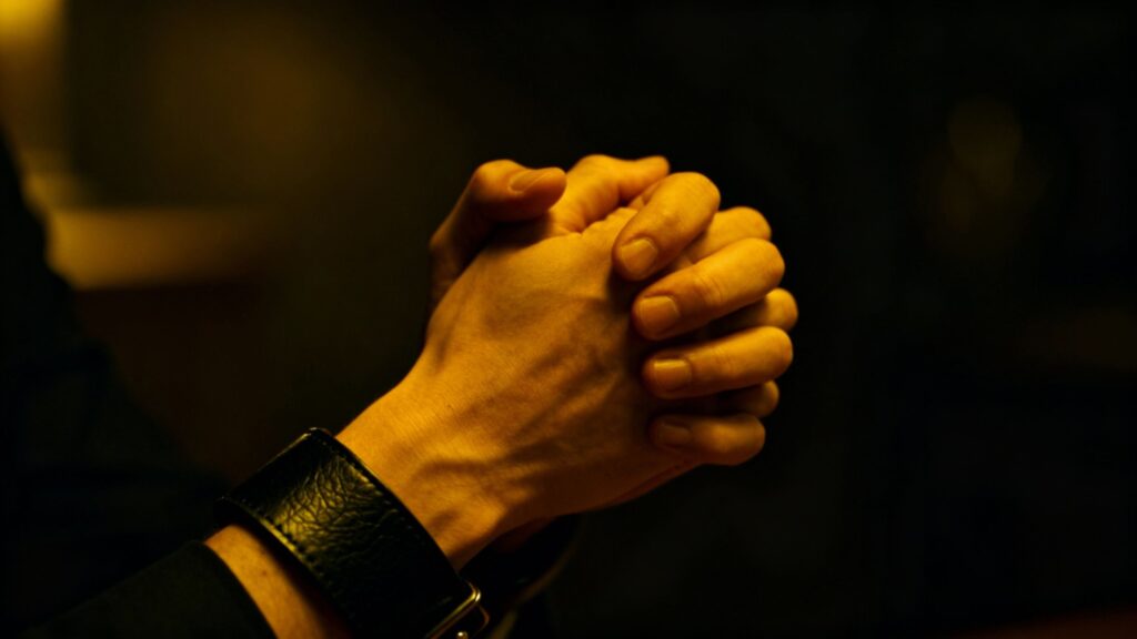Hands clasped, one with a leather cuff, in a dim setting.