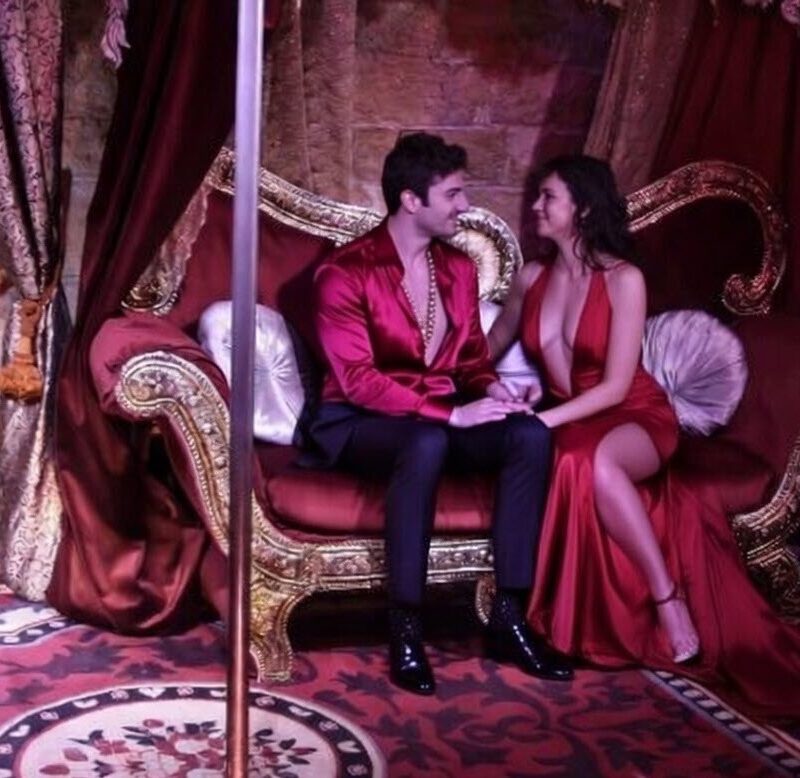 A stylish couple dressed for a night out sits closely together on an opulent, gold-trimmed red velvet chaise longue under dramatic burgundy drapery in the intimate Théâtre ChoChotte erotic theatre in Paris.